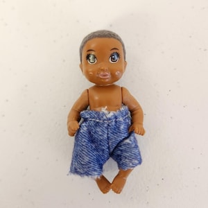 May include: A small doll with brown skin, gray hair, and blue denim shorts. The doll has large eyes and a neutral expression. The shorts are frayed at the edges. The doll is standing upright.