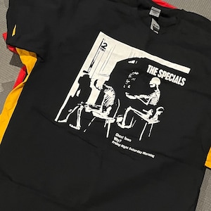 May include: Black t-shirt featuring a white graphic of a band performing, with the text "THE SPECIALS" and song titles "Ghost Town", "Why?", and "Friday Night Saturday Morning". The shirt has a V-neck and is made of a soft material.