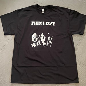 May include: Black t-shirt with a white graphic of the band Thin Lizzy. The graphic features three band members in silhouette.