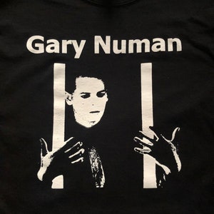 May include: Black t-shirt featuring a white graphic of Gary Numan in a prison cell. The graphic shows Numan's face and hands, with white bars behind him.