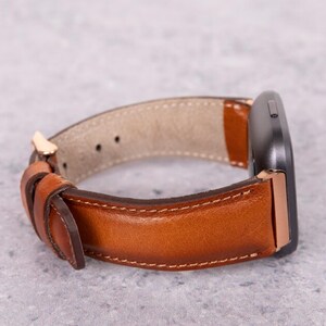 May include: A brown leather watch band with a rose gold buckle. The band is shown wrapped around a black smartwatch.