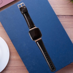 May include: A black smartwatch with a rectangular screen and a gold-coloured frame. The watch has a black leather strap with a gold buckle. The watch is on a blue surface.