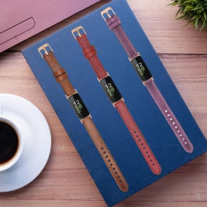 Op de afbeelding: Drie leren horlogebandjes in verschillende kleuren: bruin, rood en paars. De bandjes bevinden zich op een blauwe achtergrond en hebben gouden accenten. De bandjes zijn ontworpen voor een fitness tracker.