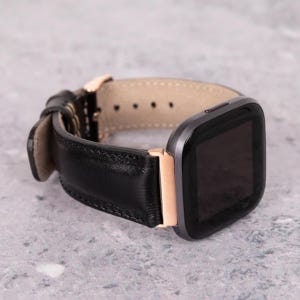 May include: Black leather watch strap with a grey watch face and gold-coloured accents. The strap is made of stitched leather and has a light tan interior. The watch face is square and has a black screen.