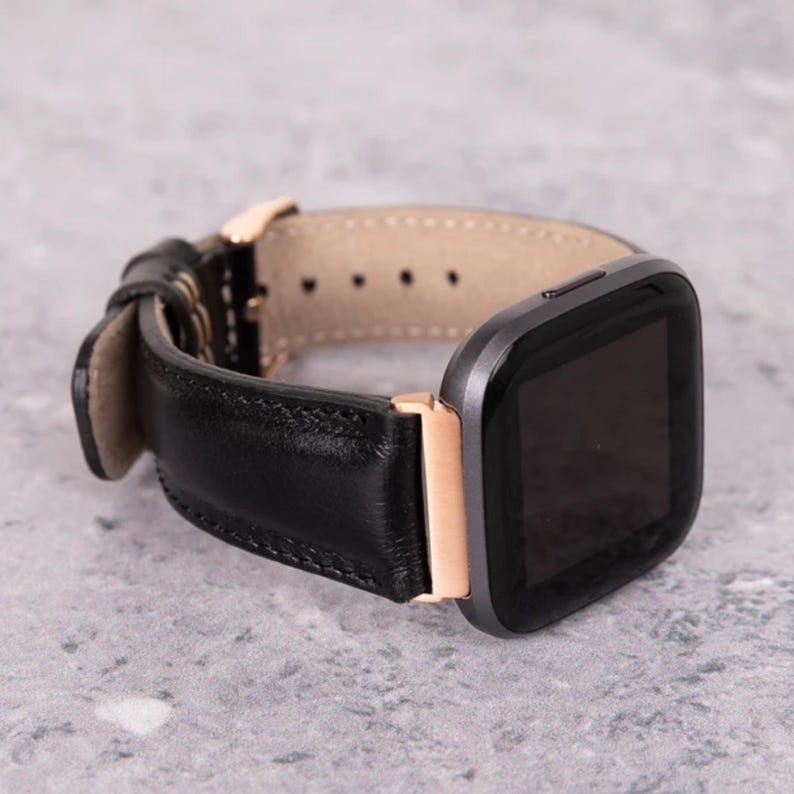 May include: Black leather watch strap with a grey watch face and gold-coloured accents. The strap is made of stitched leather and has a light tan interior. The watch face is square and has a black screen.