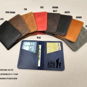 Peut inclure: Un portefeuille en cuir bleu avec deux fentes pour cartes et une poche pour l'argent. Le portefeuille porte l'inscription "SUPER DAD" et une silhouette d'un p&egrave;re et d'un enfant. Le portefeuille est pr&eacute;sent&eacute; avec d'autres portefeuilles de diff&eacute;rentes couleurs, notamment noir, marron, beige, rouge, vert et gris.