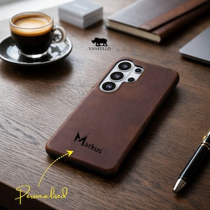 Puede incluir: Una funda de teléfono de cuero marrón con el nombre "Markus" grabado. La funda está sobre una superficie de madera, junto a una taza de café, un bolígrafo y un cuaderno. El diseño es personalizado.