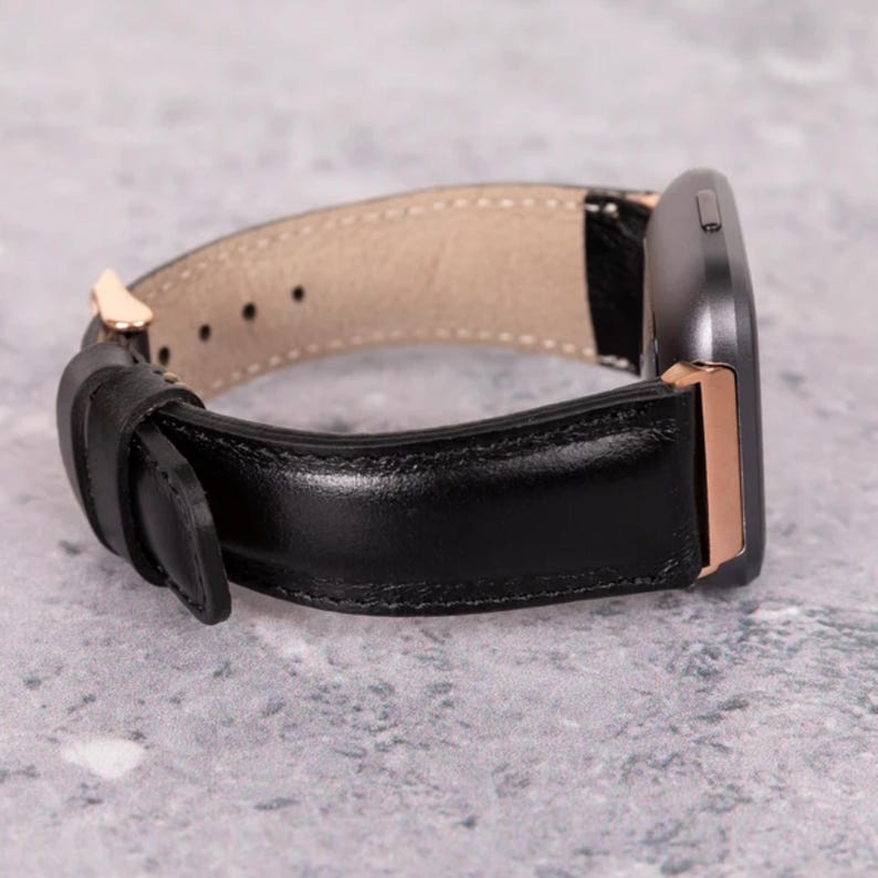 May include: Black leather watch strap with a gold-coloured buckle and a grey watch face. The strap is made of smooth, shiny leather and has a beige interior. The watch face has a rose gold-coloured frame.