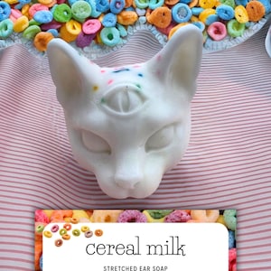 May include: White cat-shaped soap with a third eye and colorful sprinkles. The soap is labeled "cereal milk stretched ear soap with jojoba oil & mango butter".