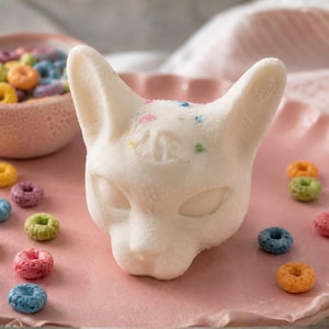 May include: A white, cat-shaped sculpture with pointed ears and almond-shaped eyes, featuring small, colorful speckles on its forehead. The sculpture is surrounded by scattered, multi-colored ring-shaped cereal pieces on a pink surface.