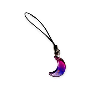 May include: A purple and pink crescent moon charm with gold flecks on a black phone strap.