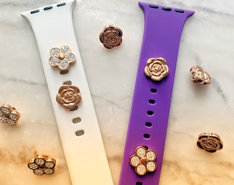 Pode incluir: Duas pulseiras para Apple Watch, uma branca e outra roxa, com pingentes decorativos de rosa e flor. Os pingentes são dourados e têm strass.