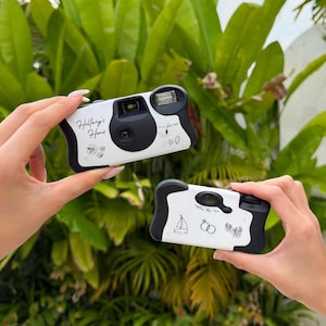 May include: Two white and black disposable cameras with custom text and illustrations. One camera reads "Hillary's Hens" with floral and star designs. The other camera has a sailboat, rings, and the phrase "capture the love."