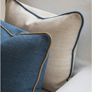 以下が含まれることがあります： 2つの装飾用クッションのクローズアップ。1つは濃い青色で茶色のトリム、もう1つは明るいベージュ色でネイビーブルーのトリムです。どちらのクッションもテクスチャード加工された生地を使用しています。