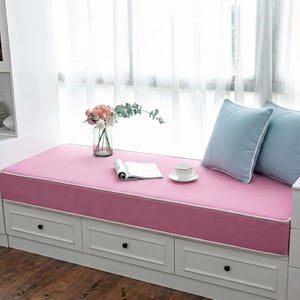 Può includere: Un cuscino rosa per la seduta della finestra con bordino bianco, una base bianca con cassetti e due cuscini azzurri. Un vaso di fiori, una tazza da tè e un libro aperto sono sul cuscino. La finestra è sullo sfondo.