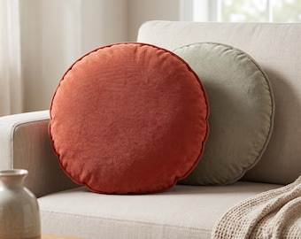 May include: Two round throw pillows with a soft, textured fabric finish. One pillow is a vibrant burnt orange color, while the second pillow positioned slightly behind it is a muted sage green. Both pillows feature a simple, circular design.