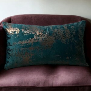 May include: Teal velvet throw pillow with gold metallic accents. The pillow has a distressed, abstract pattern.