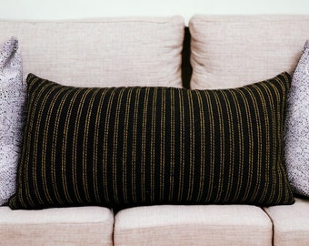 May include: A beige sofa with three cushions. The center cushion is black with thin gold stripes. The sofa is against a wood wall.
