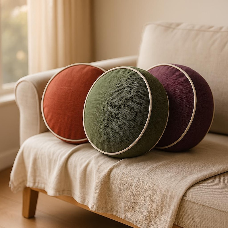 May include: Three round decorative pillows in shades of rust, olive green, and plum, each with a white trim. The pillows are arranged on a beige sofa with a matching throw. The pillows are a stylish home decor item.