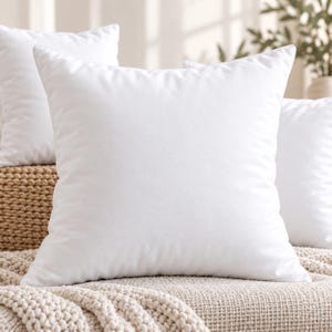 May include: A square, solid white throw pillow with a smooth, matte fabric finish. The pillow features clean, straight edges and a plump, filled appearance, presented as a singular decorative cushion for home decor and furniture arrangements.