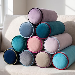 May include: A stack of ten cylindrical bolster pillows made of soft velvet fabric in various colours including lavender, teal, navy blue, light pink, grey, and beige, each featuring a contrasting piped edge around the circular end panels.