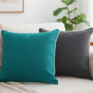 May include: Two square throw pillows on a white sofa. One pillow is teal with white piping, the other is dark gray. A beige blanket is draped over the sofa arm. Home decor.