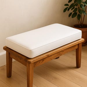 May include: A wooden bench with a rectangular white cushion. The bench has four sturdy legs and a natural wood finish. The white cushion has a contrasting white trim. The bench is suitable for indoor use.