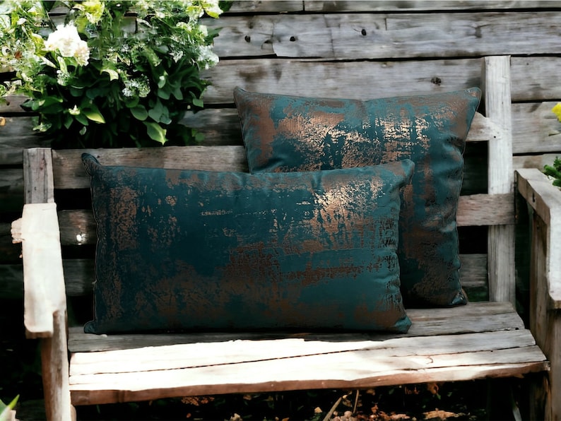 May include: Two teal green throw pillows with a copper metallic distressed pattern. The pillows are on a wooden bench.