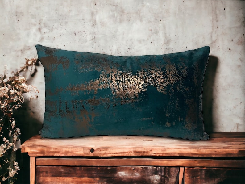 May include: A teal blue throw pillow with a gold metallic pattern. The pillow is sitting on a wooden surface.