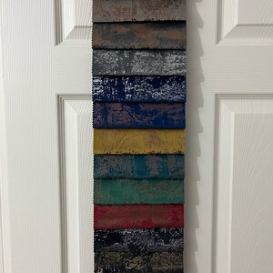 May include: A collection of 15 fabric swatches in various colors and patterns. The swatches are arranged vertically, with the top swatch being a light brown with a white distressed pattern. The swatches below are a variety of colors, including gray, blue, yellow, green, red, black, and brown. Each swatch has a different distressed pattern.