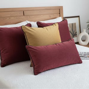 May include: A bed with a wooden headboard and white bedding. Several pillows in shades of burgundy and gold are arranged on the bed. The burgundy pillows have black trim. A magazine and decorative items are on the nightstand.