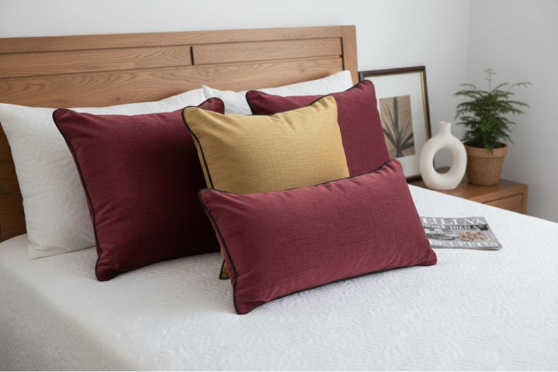 May include: A bed with a wooden headboard and white bedding. Several pillows in shades of burgundy and gold are arranged on the bed. The burgundy pillows have black trim. A magazine and decorative items are on the nightstand.