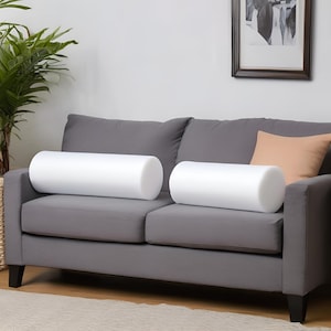 May include: Two white foam cylindrical pillows on a gray sofa with a peach colored throw pillow.