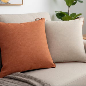 May include: Two square throw pillows on a beige sofa. One pillow is a solid burnt orange with a black trim, the other is a light beige with a brown trim. A gray blanket is draped over the sofa.