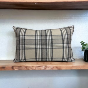 May include: A beige and brown plaid throw pillow with a rectangular shape. The pillow is sitting on a wooden shelf.