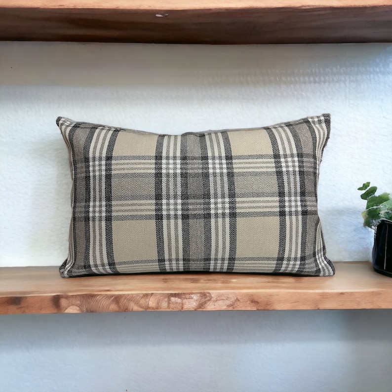 May include: A beige and brown plaid throw pillow with a rectangular shape. The pillow is sitting on a wooden shelf.
