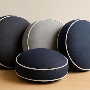 May include: Four round decorative pillows in shades of navy blue and gray, with white piping. The pillows are arranged on a wooden surface against a neutral background. The pillows vary in size and shape.