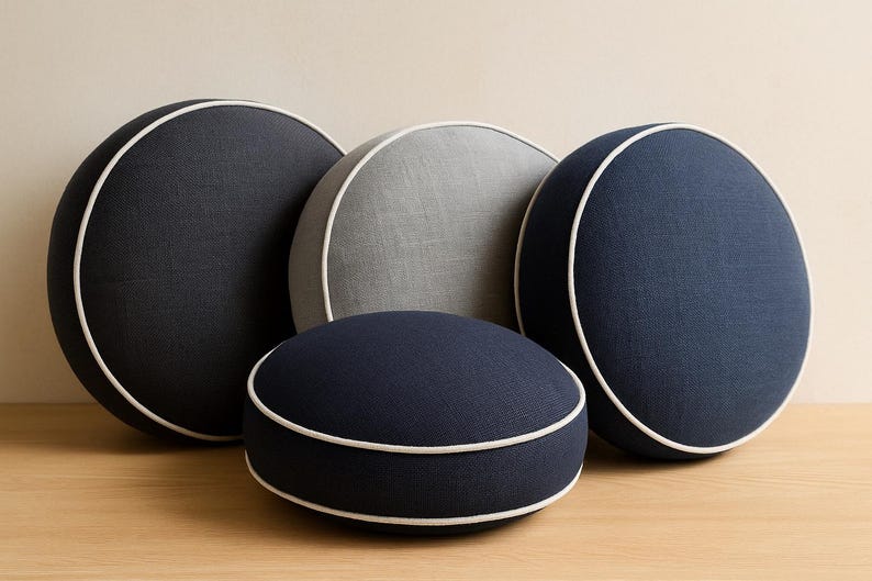 May include: Four round decorative pillows in shades of navy blue and gray, with white piping. The pillows are arranged on a wooden surface against a neutral background. The pillows vary in size and shape.