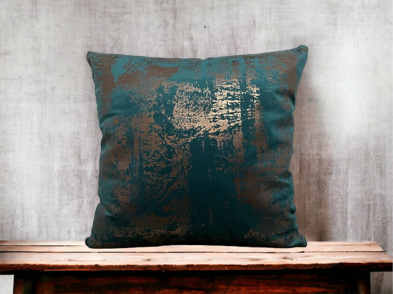 May include: Teal blue throw pillow with a gold metallic abstract design. The pillow is sitting on a wooden bench.