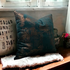 May include: A teal and gold throw pillow with a distressed pattern. The pillow is sitting on a white fluffy rug on a wooden surface.