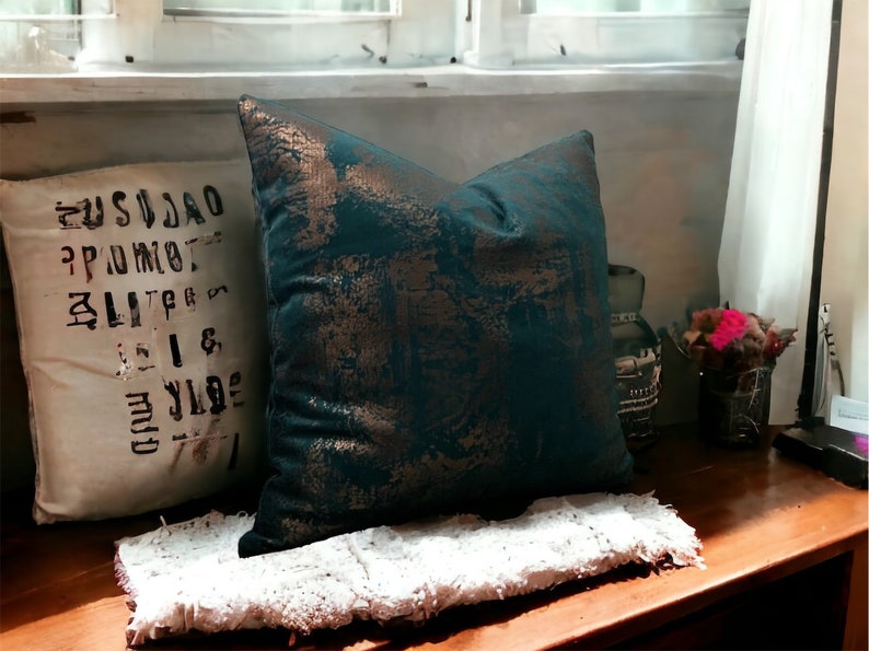 May include: A teal and gold throw pillow with a distressed pattern. The pillow is sitting on a white fluffy rug on a wooden surface.