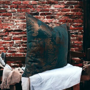 May include: A teal green throw pillow with gold accents and a distressed look. The pillow is sitting on a white surface.