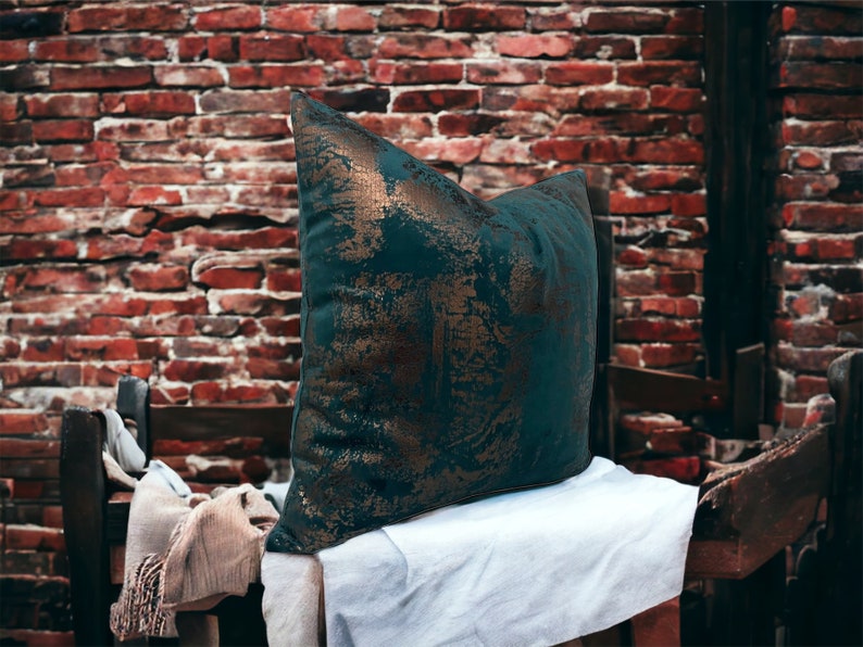 May include: A teal green throw pillow with gold accents and a distressed look. The pillow is sitting on a white surface.