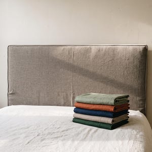 May include: A rectangular, light brown upholstered headboard with piped edges sits behind a white bed sheet. A stack of five folded rectangular textiles in sage green, burnt orange, navy blue, beige, and dark forest green rests on the bed.