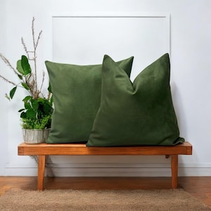 May include: Two green velvet cushion covers on a wooden bench. The cushions are square and have a soft, plush texture.