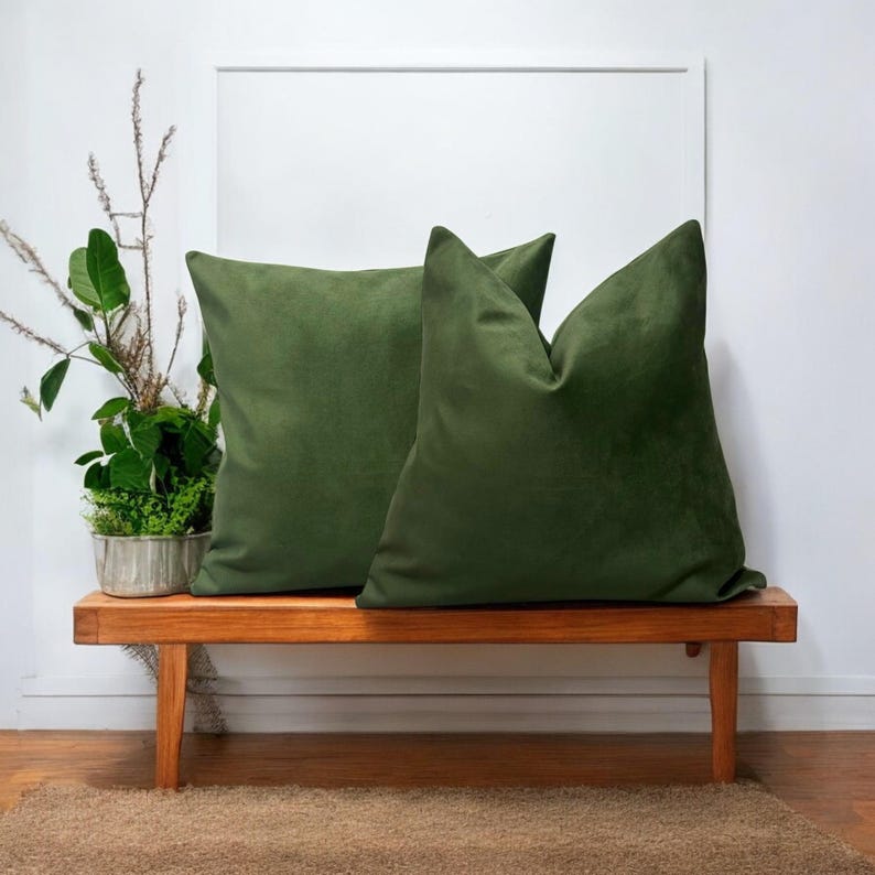 May include: Two green velvet cushion covers on a wooden bench. The cushions are square and have a soft, plush texture.