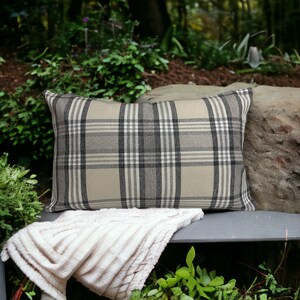 May include: A beige and black plaid throw pillow with a soft, plush texture. The pillow is sitting on a gray bench with a white blanket draped over it.