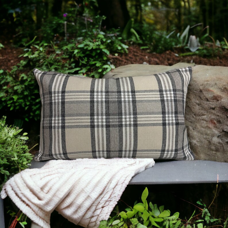 May include: A beige and black plaid throw pillow with a soft, plush texture. The pillow is sitting on a gray bench with a white blanket draped over it.