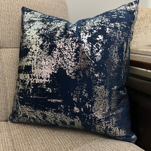 May include: A navy blue throw pillow with a silver metallic abstract pattern. The pillow is on a light beige couch.