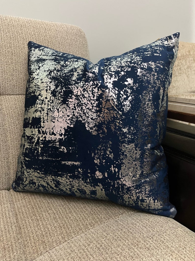 May include: A navy blue throw pillow with a silver metallic abstract pattern. The pillow is on a light beige couch.